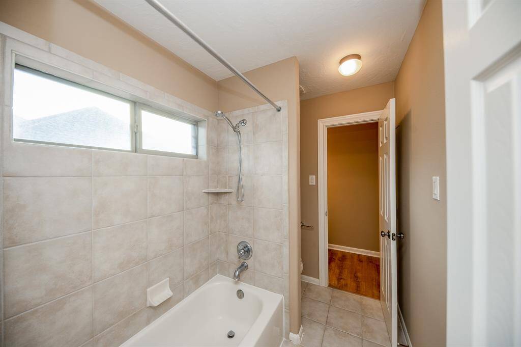 This bathroom features a clean, tiled bathtub and shower area with a high window for natural light. The neutral walls and ceiling light create a warm atmosphere, complemented by tile flooring.