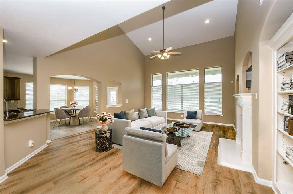 The living area features high ceilings, large windows for natural light, and an open layout connecting to the dining space. Warm colore d flooring and neutral tones create a welcoming atmosphere. A ceiling fan and built-in shelves add functionality and style. Image is Virtually Staged.