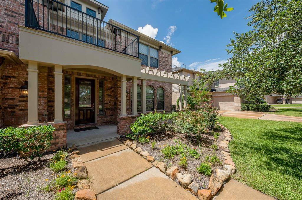 This inviting home features a charming brick exterior with a welcoming front porch and pergola. The well-maintained landscaping includes a pathway lined with stone borders, leading to the front door. The property boasts a spacious, lush lawn, offering great curb appeal in a pleasant neighborhood setting.