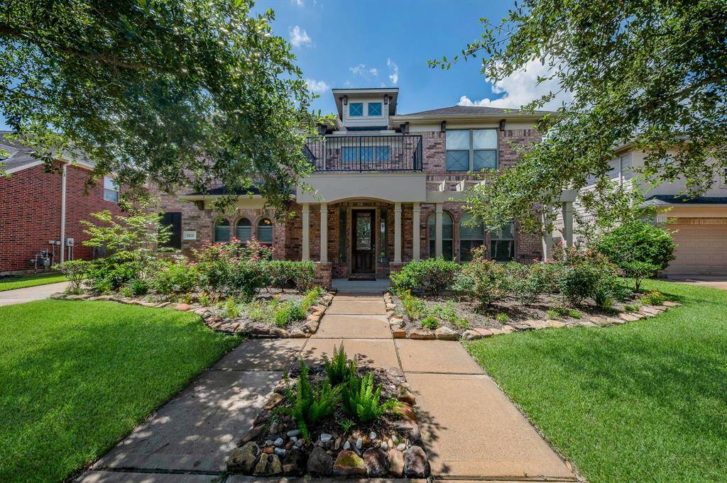 This photo showcases a charming two-story brick home with a welcoming front porch and balcony. The landscaped front yard features lush greenery, a neat pathway, and vibrant flower beds, offering great curb appeal.