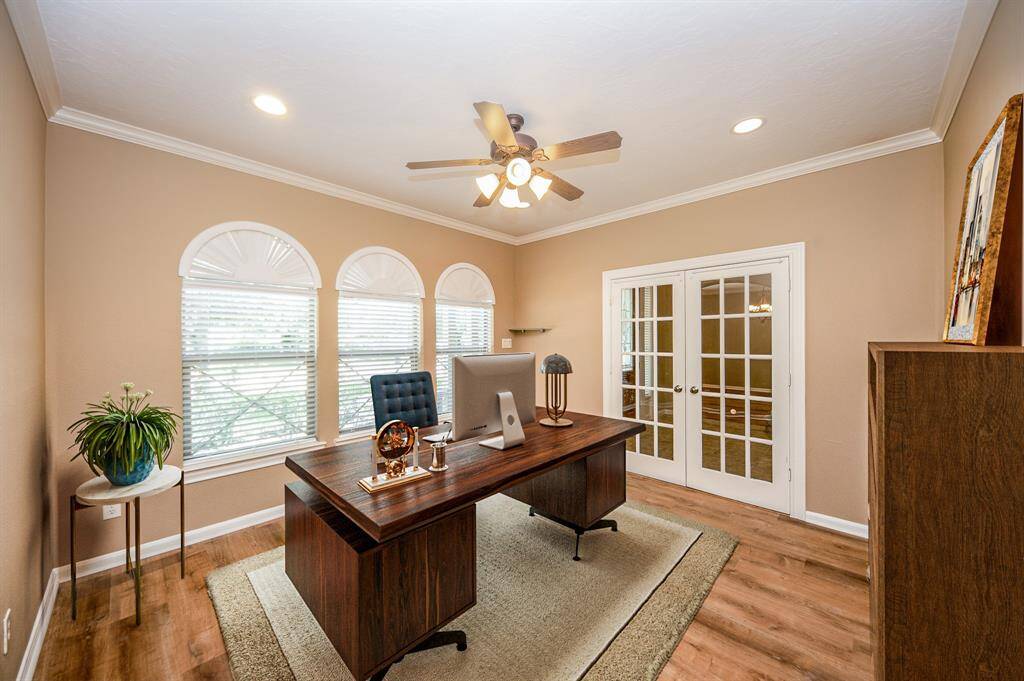 This photo showcases a bright home office with a large wooden desk, ample natural light from three arched windows, and elegant French doors. The room features recessed lighting, a ceiling fan, and hardwood flooring, offering a cozy yet professional atmosphere.  Image is Virtually Staged.