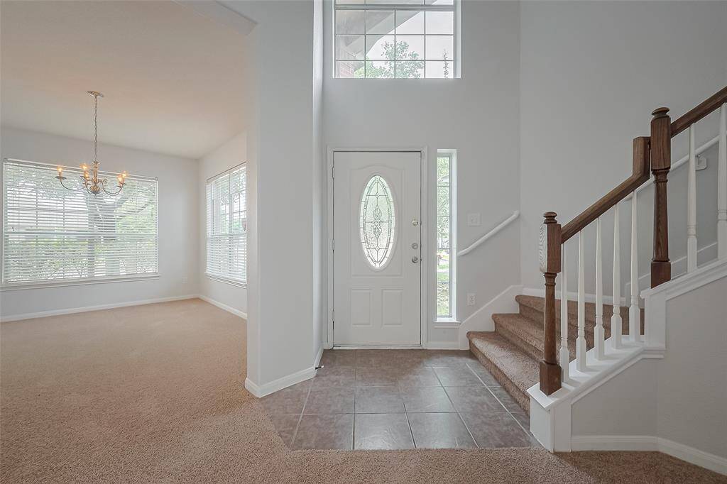 This home features a bright entryway with a decorative front door, high ceiling, and a staircase with wooden railings. To the left, an inviting formal dining area with large windows and a chandelier offers ample natural light. The space blends tiled and carpeted flooring.