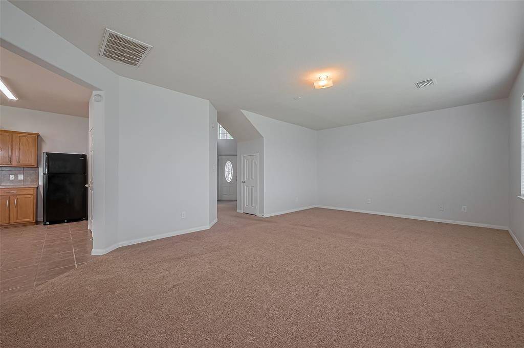 Spacious living area with neutral walls and carpet, featuring a bright ceiling light. It opens to a kitchen with wooden cabinets and a black refrigerator, and has ample natural light from multiple windows.