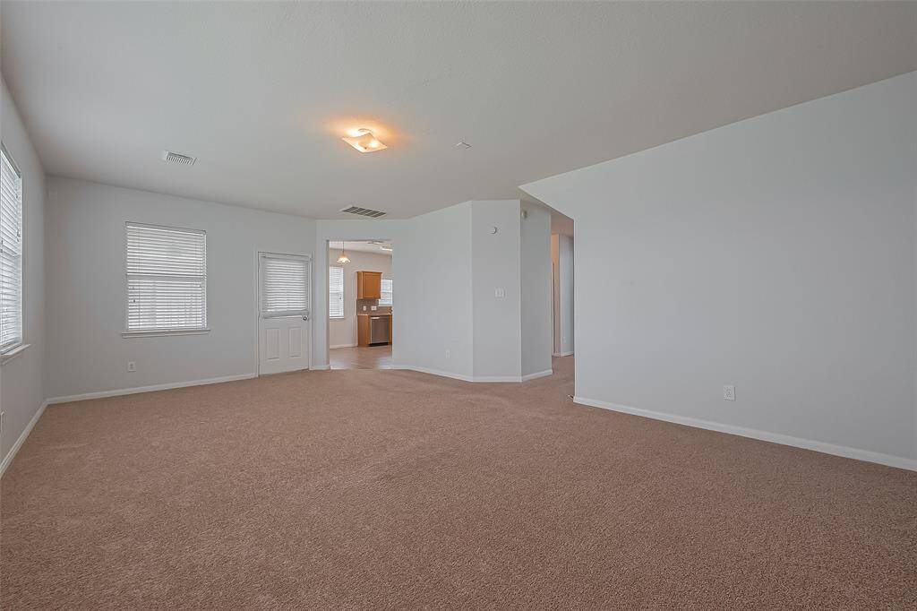 Spacious living area with neutral carpet and walls, featuring ample natural light from multiple windows. The room flows into an adjacent kitchen area, providing an open-concept feel.
