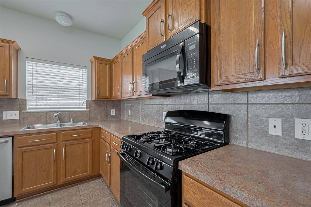 This kitchen features modern appliances, including a gas stove and microwave, complemented by wooden cabinetry and tiled countertops. Natural light enters through a window above the sink, enhancing the space.