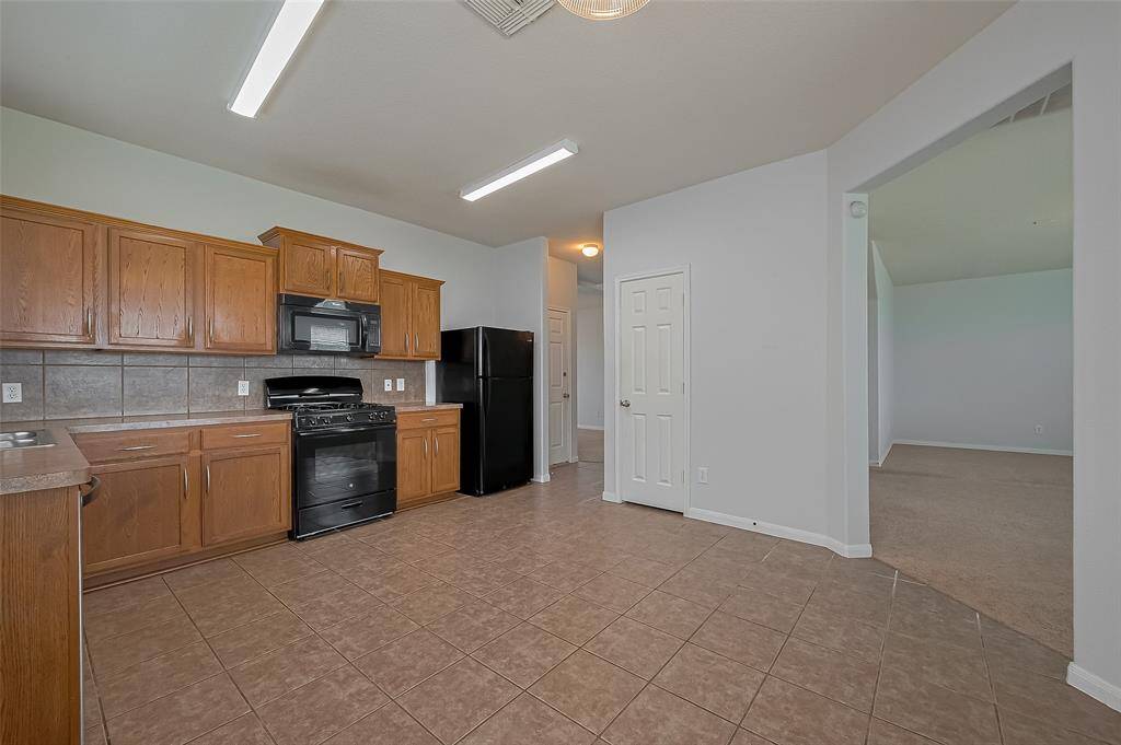 This photo shows a spacious kitchen with wooden cabinets, a black stove, and a refrigerator. It has tiled flooring and neutral walls, leading to an adjacent carpeted room.