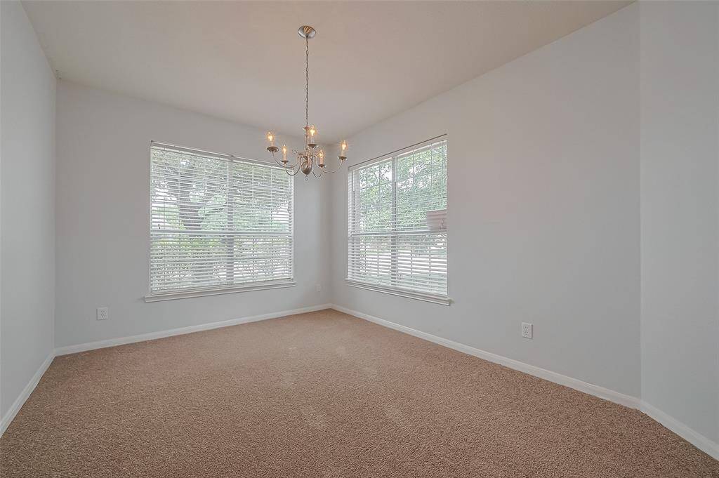 This room features neutral carpeting, light gray walls, and a modern chandelier. Large windows provide ample natural light and offer views of greenery outside. Ideal for a dining or sitting area.