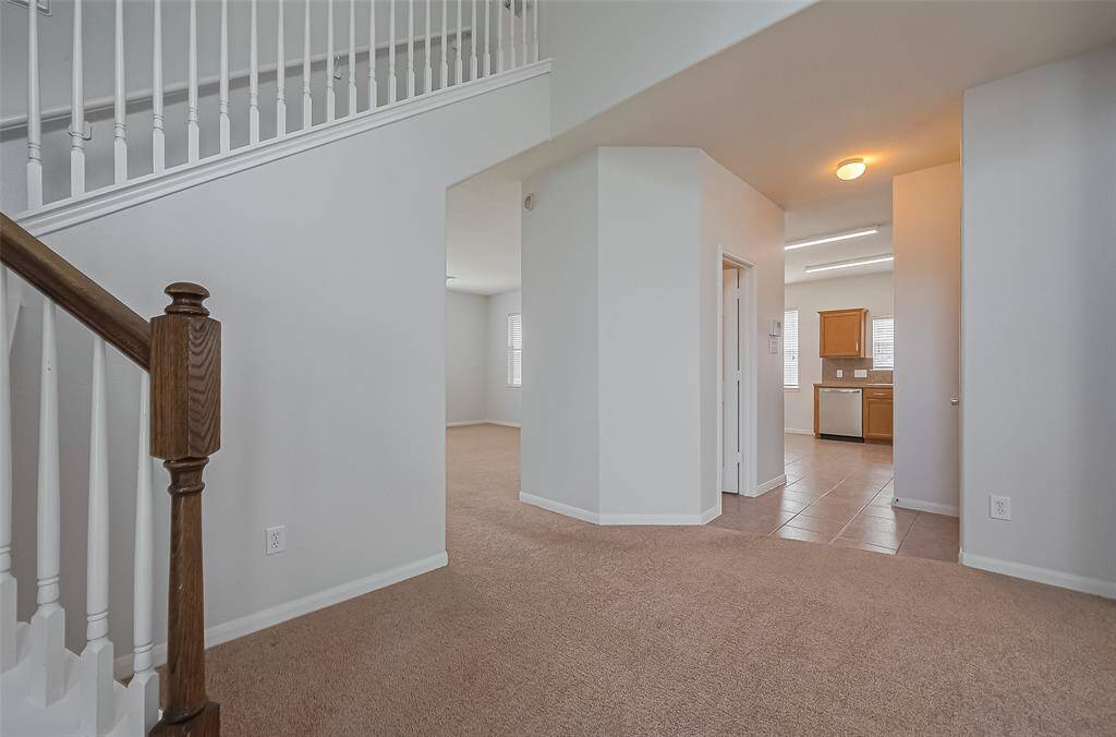 The photo shows an inviting open-plan entryway with a carpeted floor leading to a kitchen with tile flooring and wooden cabinetry. A staircase with white railings and a wooden banister adds charm and character. The space is well-lit, creating a warm and welcoming atmosphere.