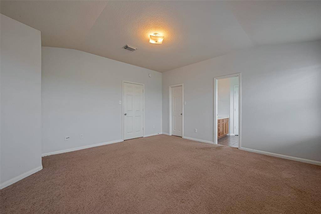 Spacious bedroom with neutral carpet and light-colored walls, featuring a ceiling light fixture. It has a door leading to an en-suite bathroom with visible cabinetry.