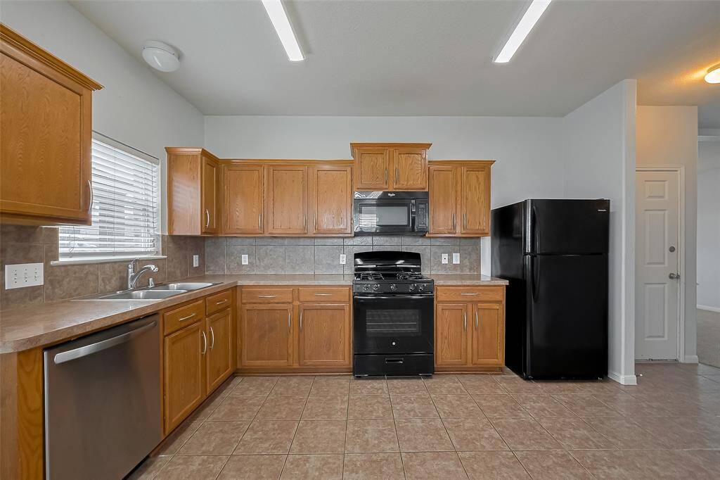 This kitchen features wood cabinetry, a mix of black and stainless steel appliances, tiled flooring, and ample counter space. It also has a window for natural light and a functional layout.