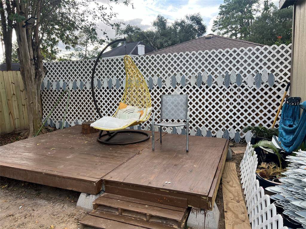 This photo shows a cozy outdoor patio area with tiled flooring and a small table with chairs. There are several potted plants adding greenery, and a wooden fence provides privacy. A door leads inside, and a parked vehicle is visible through an open gate.