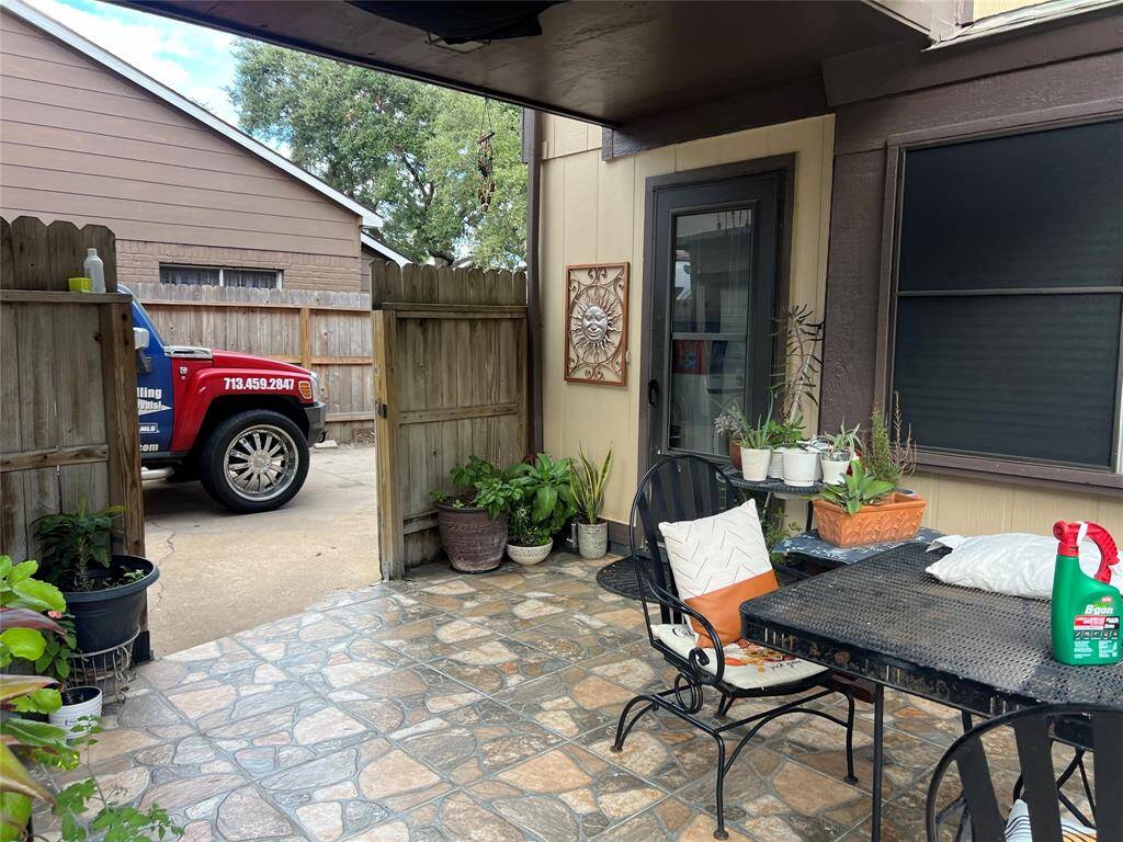 Charming patio area with decorative tile flooring, surrounded by potted plants. Features outdoor seating and a view of a wooden gate leading to the driveway. Cozy and inviting for relaxation or entertaining.