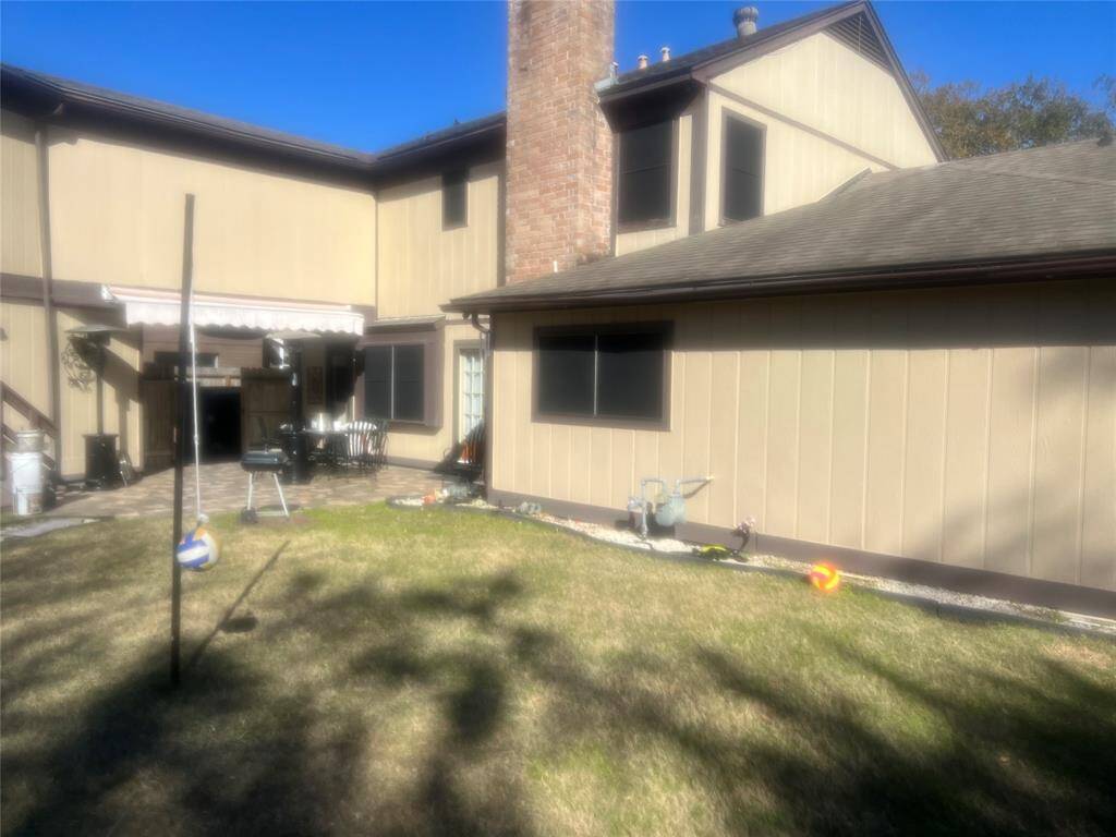This photo shows a backyard with a grassy area and a patio. There's an outdoor seating arrangement and a barbecue grill. The house exterior is beige with a brick chimney, and there's a shade awning over the patio.