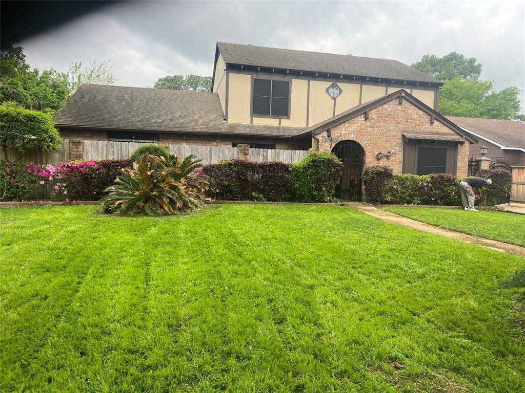This photo shows a two-story home with a mix of brick and siding exteriors. It features a well-maintained front yard with lush green grass, a variety of shrubs, and vibrant flowering plants. The property is enclosed by a wooden fence, offering privacy and charm.