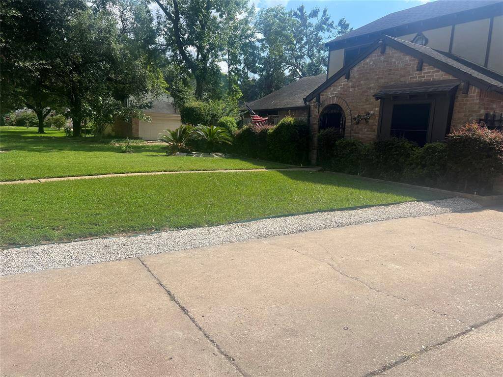 Charming brick home with a lush, green front lawn and mature trees. The driveway has a gravel border, and the house features large windows and a well-maintained garden. Peaceful suburban setting.