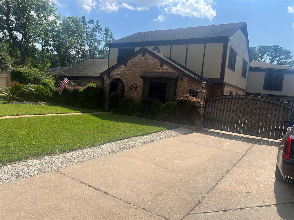 This photo shows a two-story brick house with a well-maintained lawn and a driveway leading to a gated entrance. The home features a combination of brick and siding, with mature trees and manicured shrubs in the front yard, creating a welcoming and private atmosphere.