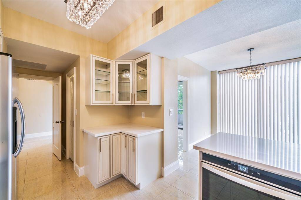 This kitchen features modern stainless steel appliances, elegant glass-front cabinets, and a spacious layout. It includes a chic chandelier, ample natural light from large windows with vertical blinds, and a sleek countertop, perfect for dining and entertaining.