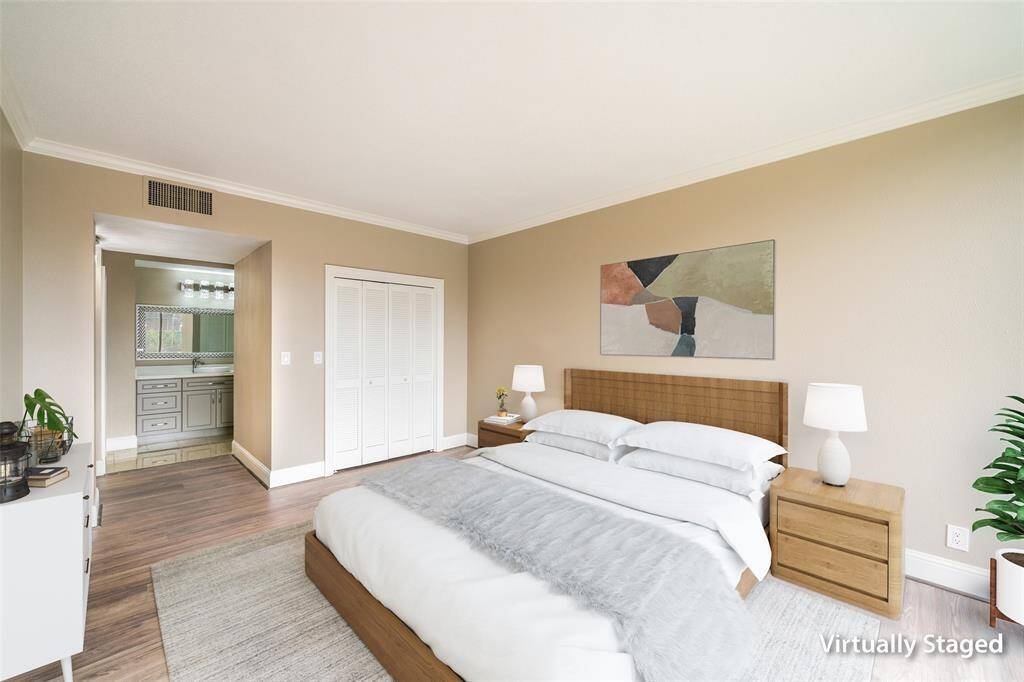 This is a virtually staged bedroom featuring neutral walls, wood flooring, and ample natural light. A doorway leads to a bathroom with a sleek vanity.