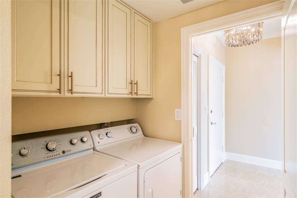 This photo shows a bright laundry room featuring a washer and dryer, overhead storage cabinets, and a modern chandelier, with a tiled floor and a doorway leading to another room.