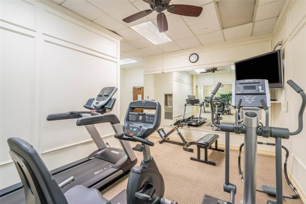 This gym features modern exercise equipment, including treadmills and an elliptical. The room is well-lit with a ceiling fan, mirrored wall, and a mounted TV for convenience. Ideal for fitness enthusiasts.