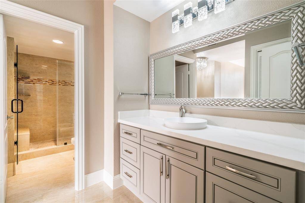 This bathroom features a modern design with a large vanity, sleek countertop, and stylish round sink. There's a wide mirror with elegant detailing above. The spacious walk-in shower has glass doors and tasteful tile accents, adding a touch of luxury.