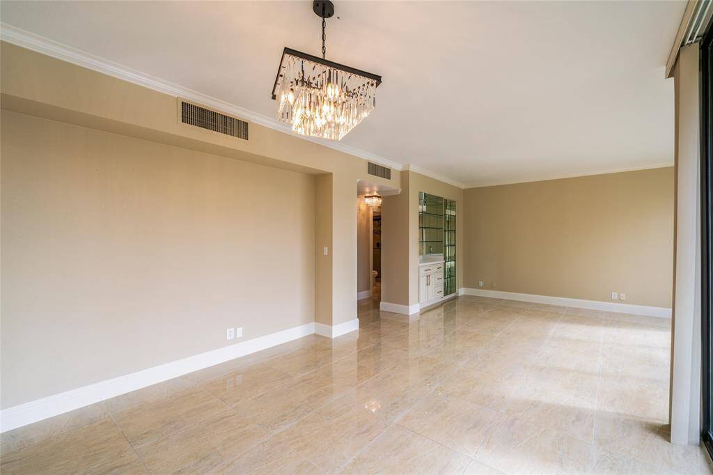 The room is spacious with glossy tiled floors and neutral walls, featuring a modern chandelier. There's built-in shelving with mirrored backing and ample natural light from large windows, creating a bright and inviting space.