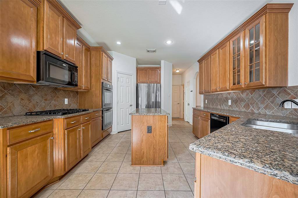 This spacious kitchen features elegant wooden cabinetry, granite countertops, and a central island. It includes modern appliances, a gas cooktop, and ample storage, complemented by tiled flooring and a stylish backsplash. Ideal for culinary enthusiasts and gatherings.