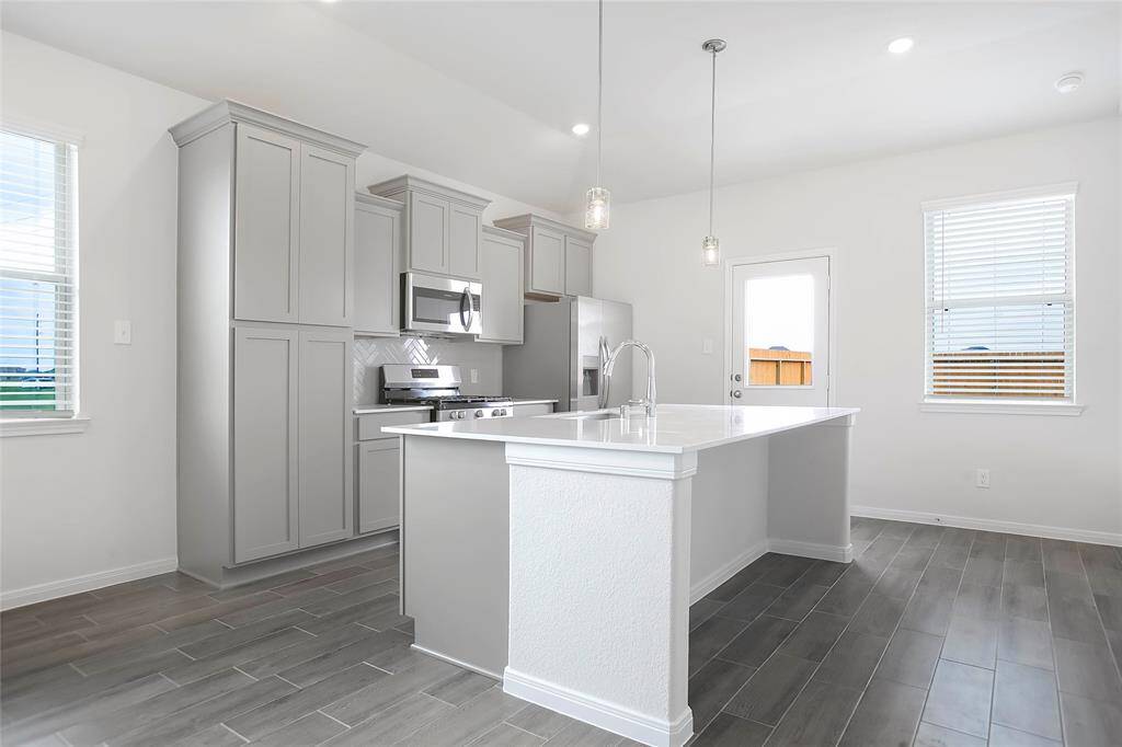 This spacious kitchen features large windows, high ceilings, wood cabinets, granite countertops, modern tile backsplash, recessed lighting with additional custom pendant lighting, extended counter space, granite kitchen island with sink and space for breakfast bar.