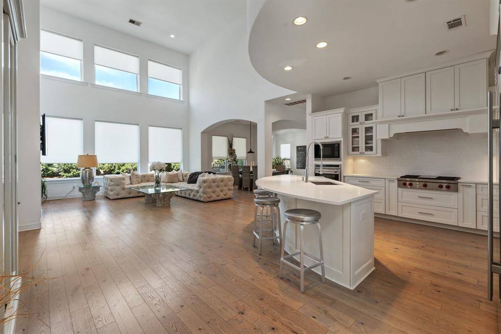 This spacious open-concept living area features high ceilings and large windows for abundant natural light. The modern kitchen includes a central island, sleek white cabinetry, and high-end appliances. The adjoining living room offers a cozy seating area.