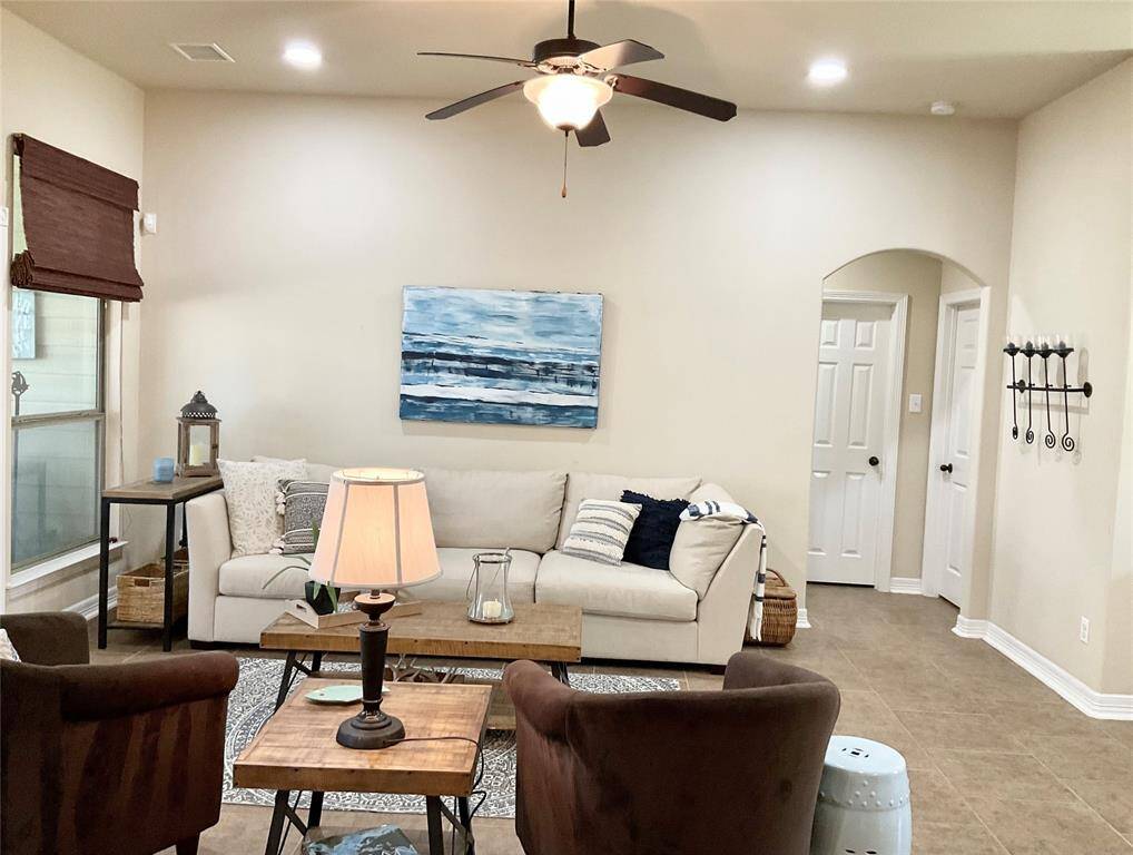 The inviting living room features a high ceiling with a ceiling fan, neutral walls, and tiled flooring. Natural light streams through large windows by the entrance, enhancing the cozy seating area.