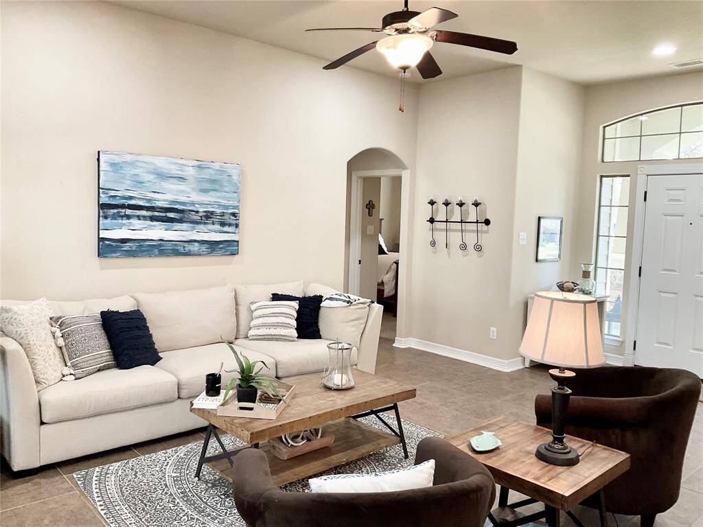 The inviting living room features a high ceiling with a ceiling fan, neutral walls, and tiled flooring. Natural light streams through large windows by the entrance, enhancing the cozy seating area.