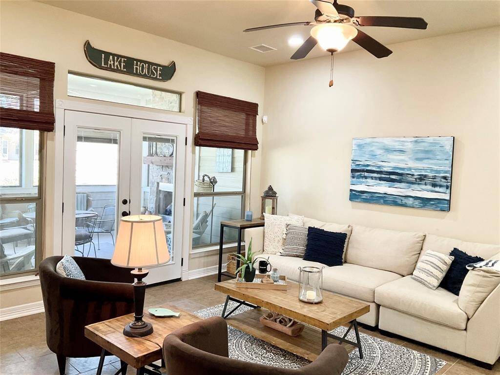 The inviting living room features a high ceiling with a ceiling fan, neutral walls, and tiled flooring. Natural light streams through large windows by the entrance, enhancing the cozy seating area.
