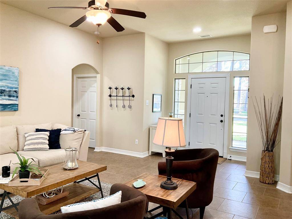 The inviting living room features a high ceiling with a ceiling fan, neutral walls, and tiled flooring. Natural light streams through large windows by the entrance, enhancing the cozy seating area.