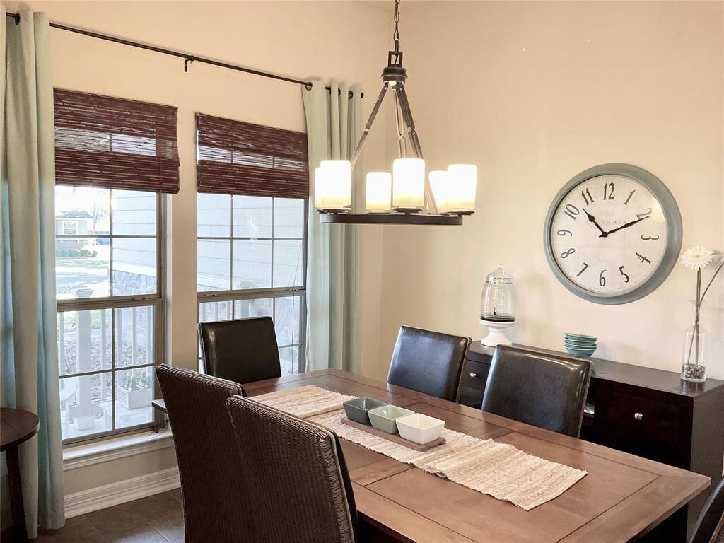 The dining room features a warm, inviting atmosphere with a wooden table and dark chairs. It has large windows with bamboo shades, a modern chandelier.   The neutral wall color pairs well with light blue curtains, creating a cozy space for meals.