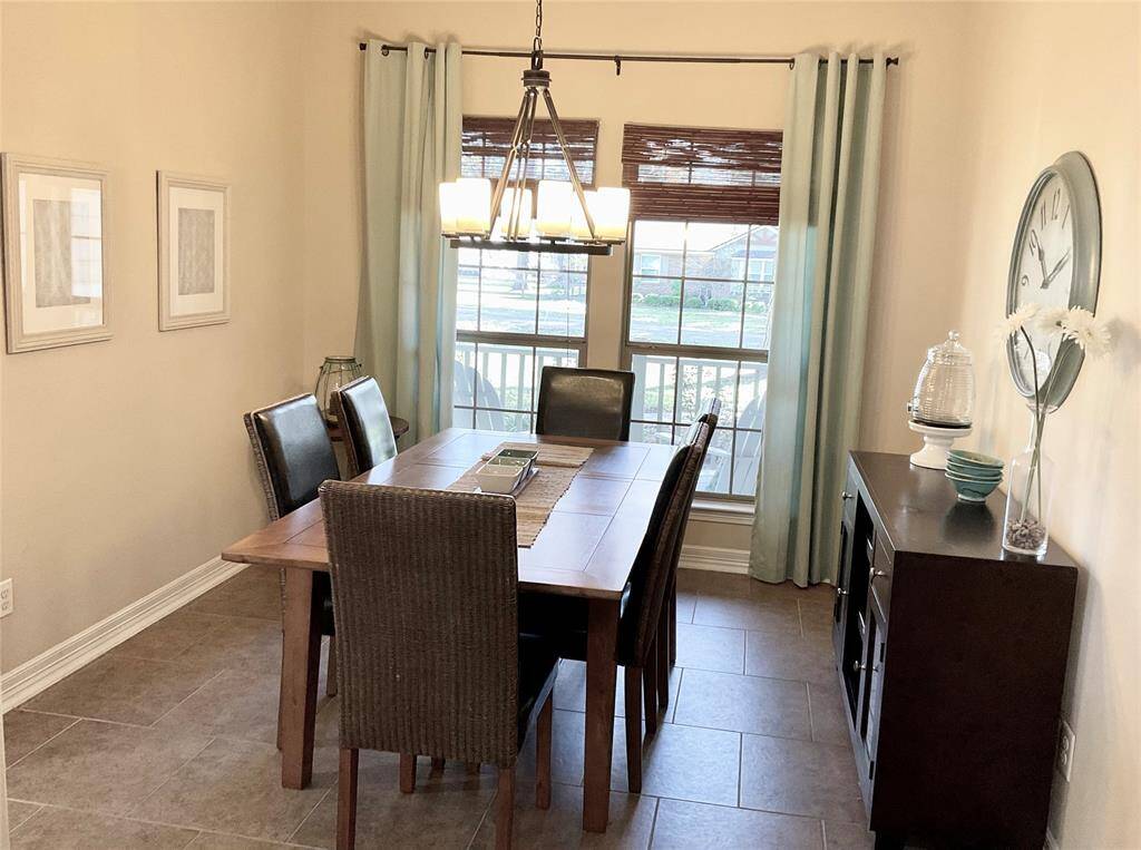 The dining room features a warm, inviting atmosphere with a wooden table and dark chairs. It has large windows with bamboo shades, a modern chandelier.   The neutral wall color pairs well with light blue curtains, creating a cozy space for meals.