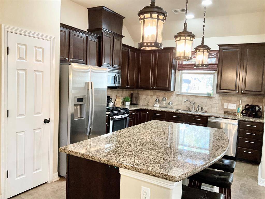 This kitchen features dark wooden cabinetry, granite countertops, and stainless steel appliances, including a fridge and dishwasher. It has a large island with seating and stylish hanging lights, creating a warm, inviting space.  The heart of the home.