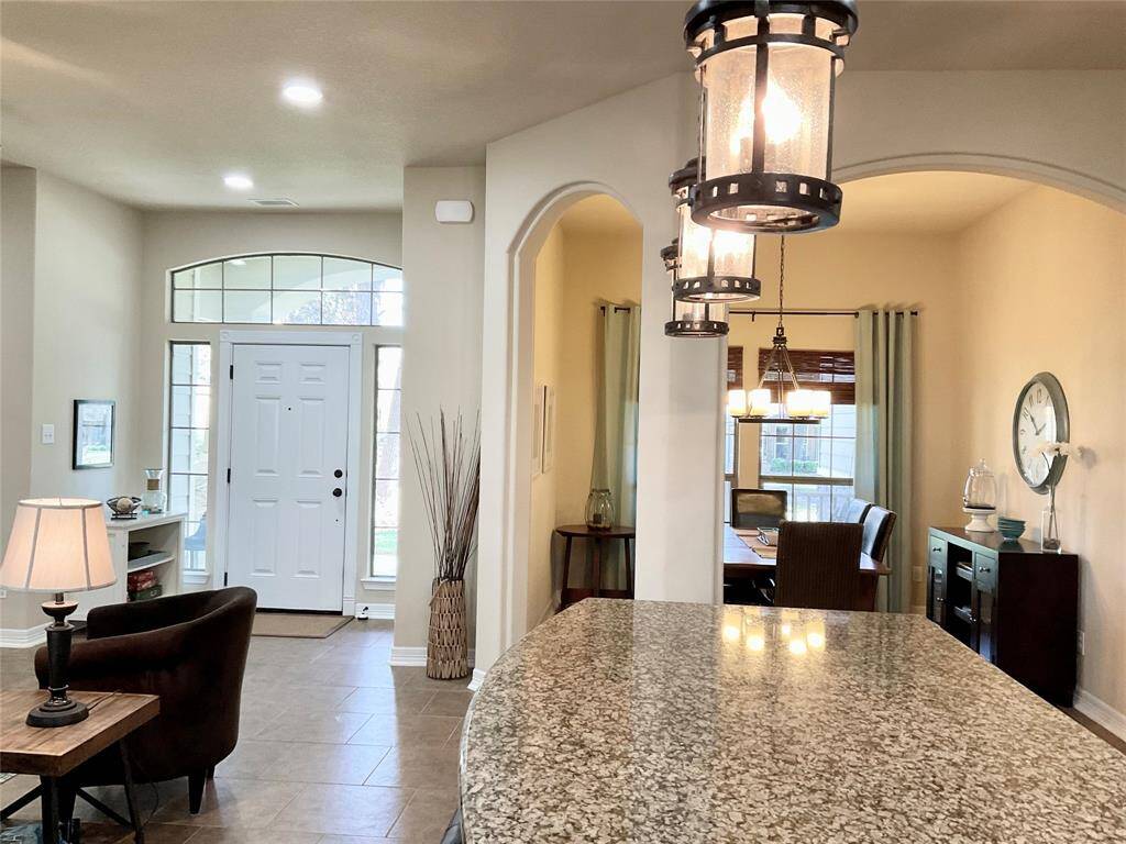 Enjoy a welcoming entryway with a white front door and large windows, allowing natural light to fill the space. The open floor plan includes a view of a dining area with stylish lighting. The interior features neutral tones and a granite countertop, creating a modern and inviting atmosphere.