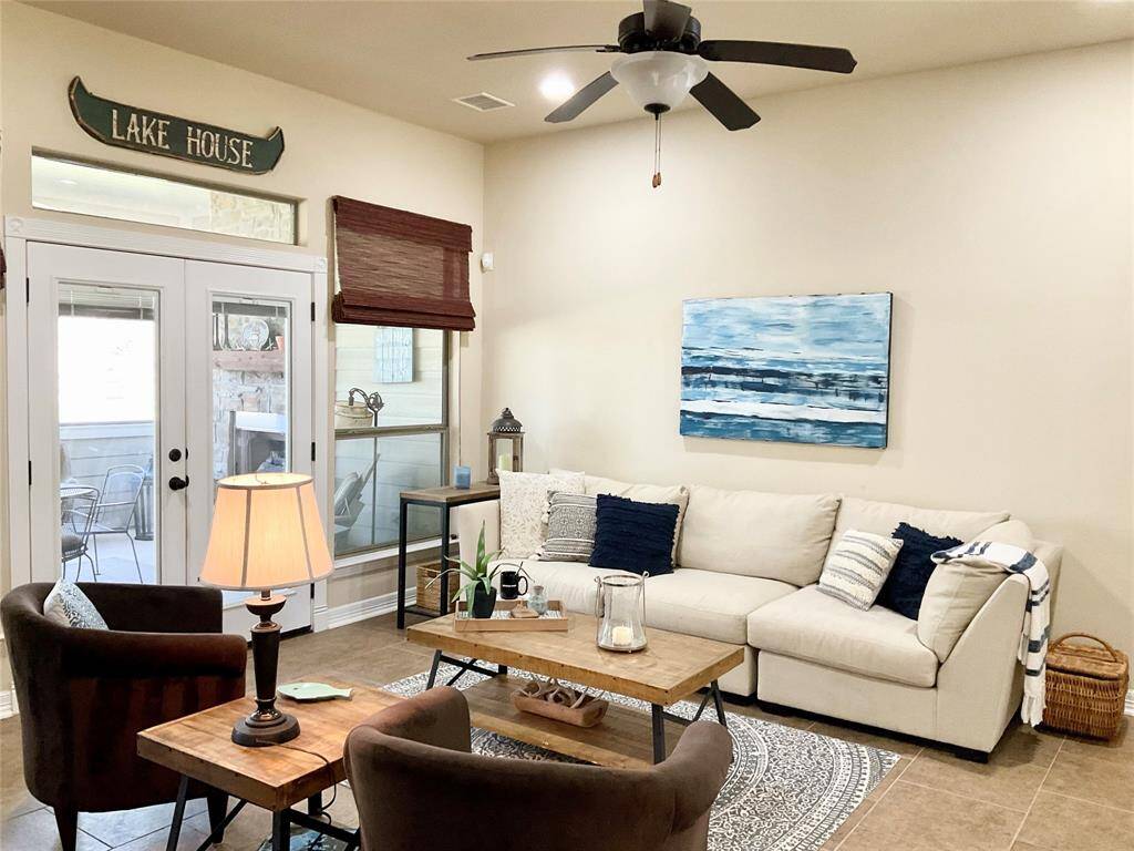 The inviting living room features a high ceiling with a ceiling fan, neutral walls, and tiled flooring. Natural light streams through large windows by the entrance, enhancing the cozy seating area.