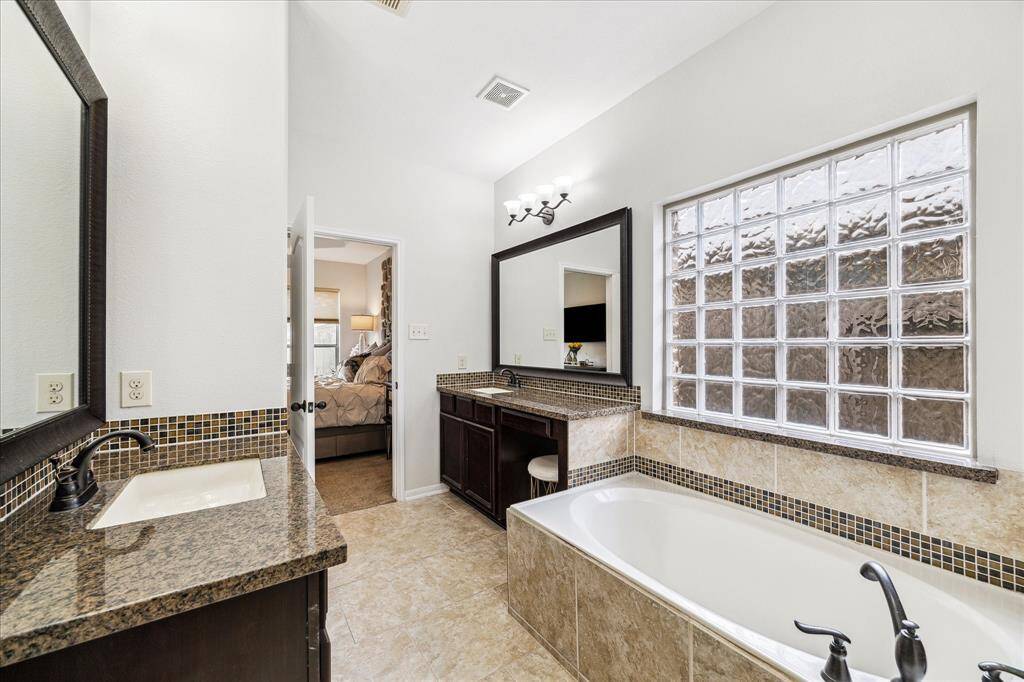 This bathroom features dual granite vanities, a large soaker tub with modern fixtures, and a glass block window providing natural light.