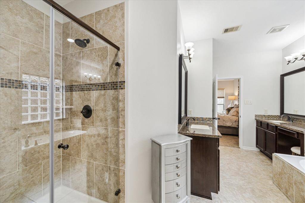 This bathroom features a spacious walk-in shower with elegant tile work, dual vanity sinks with dark wood cabinets, and a glimpse into the Primary bedroom.