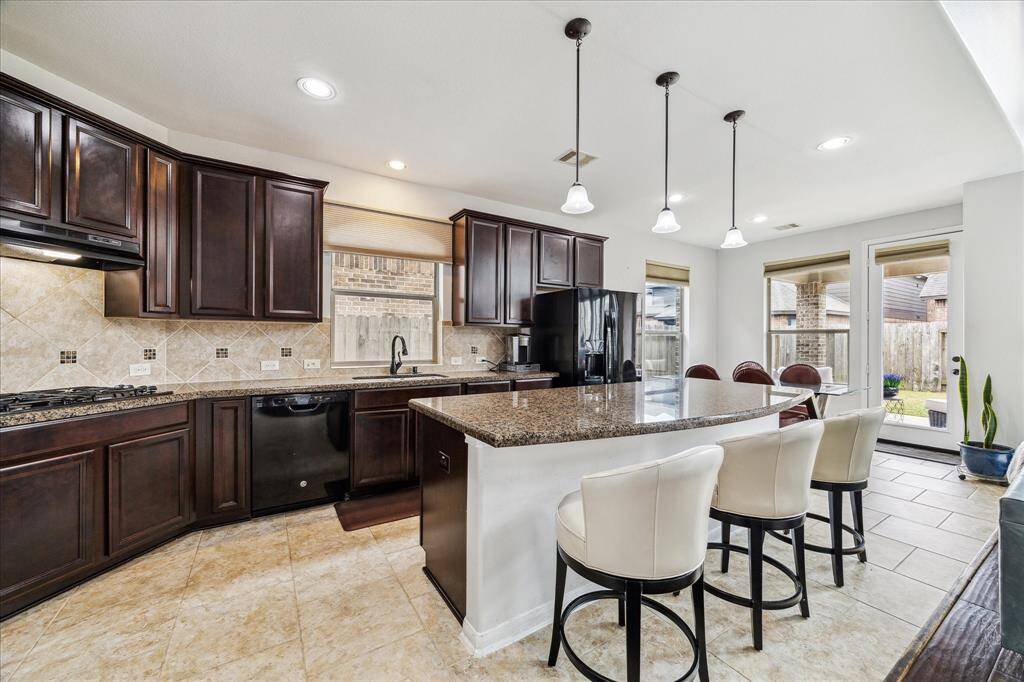 This modern kitchen features dark wood cabinetry, granite countertops, and a large island with seating. It includes tile flooring, pendant lighting, and ample natural light from large windows and glass doors leading to the covered patio and  backyard.