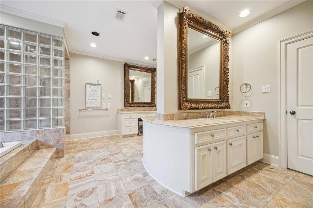 The serene primary bedroom is a luxurious retreat, complete with French doors that open to a private terrace, offering seamless indoor- outdoor living. A gaslog fireplace with a wood mantle and tile surround creates a cozy focal point, while a wall of windows provides picturesque views of the back lawn. Custom designer window treatments, recessed LED lighting, and custom crown molding enhance the room’s elegant atmosphere.
