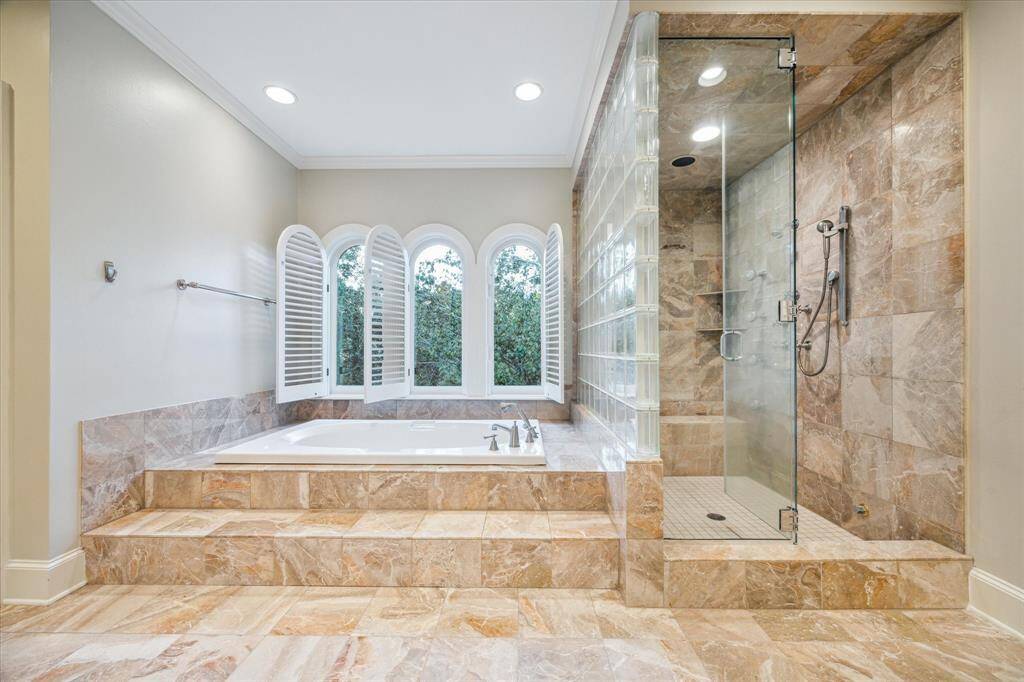The luxurious primary bathroom features stunning marble tile flooring and countertops, with dual vanities and Perrin and Rowe polished nickel faucets adding an elegant touch. A Passport Ensemble jetted tub and walk-in shower with steam function offer spa-like amenities, while custom crown molding and recessed LED lighting enhance the space’s refined atmosphere.