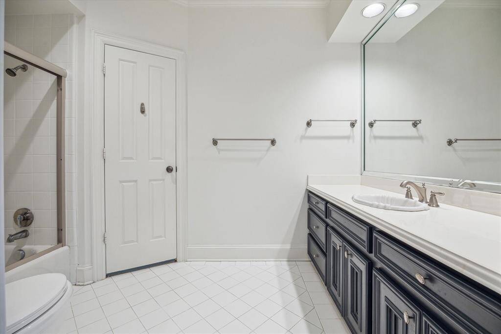 The ensuite bathroom features a custom beveled mirror, sleek Corian countertops with a brushed nickel widespread faucet, and a pristine white sink. The tub/shower combo with a custom glass enclosure and recessed LED lighting adds both functionality and style, while designer neutral wall paint complements the space’s sophisticated design. Access to a spacious walk-in closet with built-in drawers, hanging space, and open shelving makes this bathroom as practical as it is luxurious.