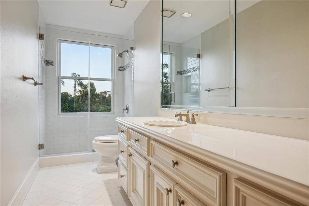 This luxurious full bathroom features a custom beveled mirror, Corian countertops with a brushed nickel widespread faucet, and a pristine white sink, offering both style and functionality. The walk-in shower, with its custom glass enclosure and a window providing western exposures, creates a serene space, while recessed LED lighting illuminates the designer neutral wall paint and white tile flooring.