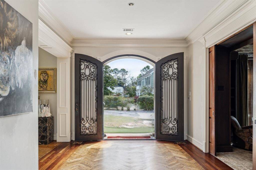 The grand entry of this luxury home is defined by warm hardwood flooring with a stunning custom herringbone inlay and surround detail. A custom steel and glass French door makes a striking first impression, flanked by dual coat closets for convenience. Recessed LED lighting and custom crown molding accentuate the designer neutral wall paint, creating an elegant and welcoming space.