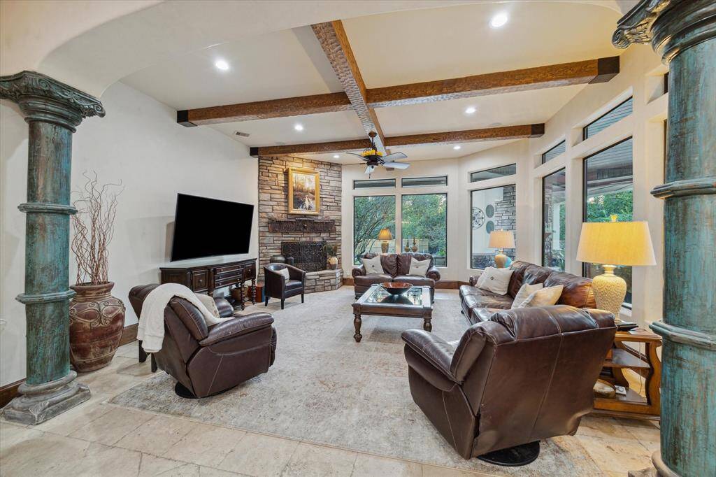 Spacious living room featuring high ceilings with exposed wooden beams, large windows for ample natural light, a stone fireplace, and elegant columns. The room is furnished with comfortable leather seating and has a warm, inviting ambiance.