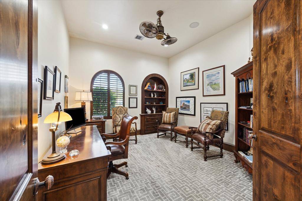 This is a carefully planned and well appointed home office featuring rich wood furnishings, a built-in bookshelf, a large arched window with plantation shutters, and a classic desk. The room has a high ceiling with a traditional ceiling fan and is decorated with framed artwork, providing a sophisticated and comfortable workspace.