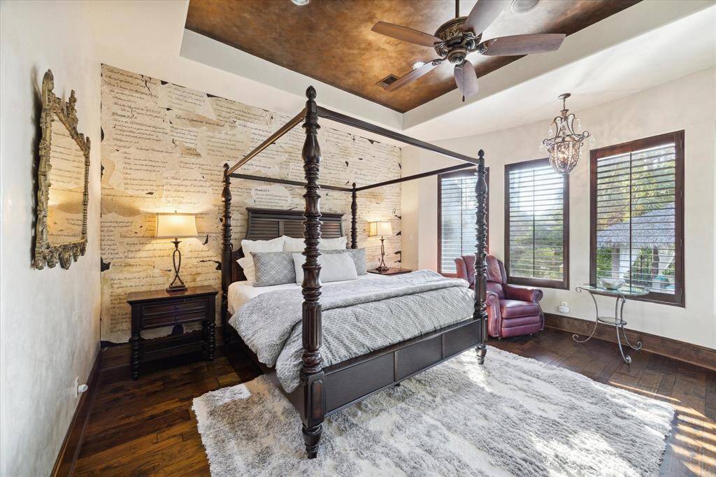 This is the elegant primary bedroom featuring  hardwood floors, a unique script-patterned accent wall, and large windows allowing for ample natural light. The room includes a comfortable seating area, stylish lighting fixtures, and a warm, inviting color palette. It is privately tucked away.