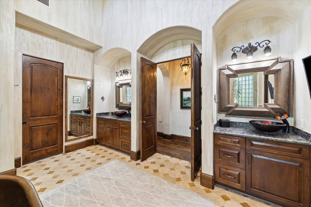 Primary custom bathroom with elegant wooden cabinetry, a dark marble countertop, and a unique vessel sink. The space features arched doorways, a large framed mirror, and a blend of tiled and hardwood flooring, creating a sophisticated and warm atmosphere.